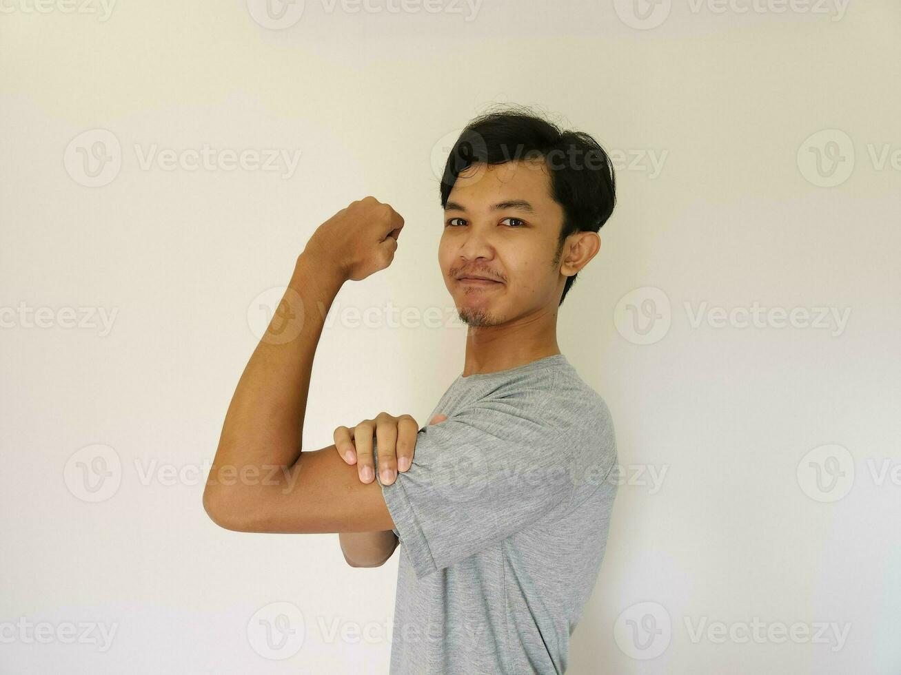 aufgeregt asiatisch Mann tragen ein grau Hemd zeigen stark Geste durch Heben seine Waffen und Muskeln lächelnd stolz foto