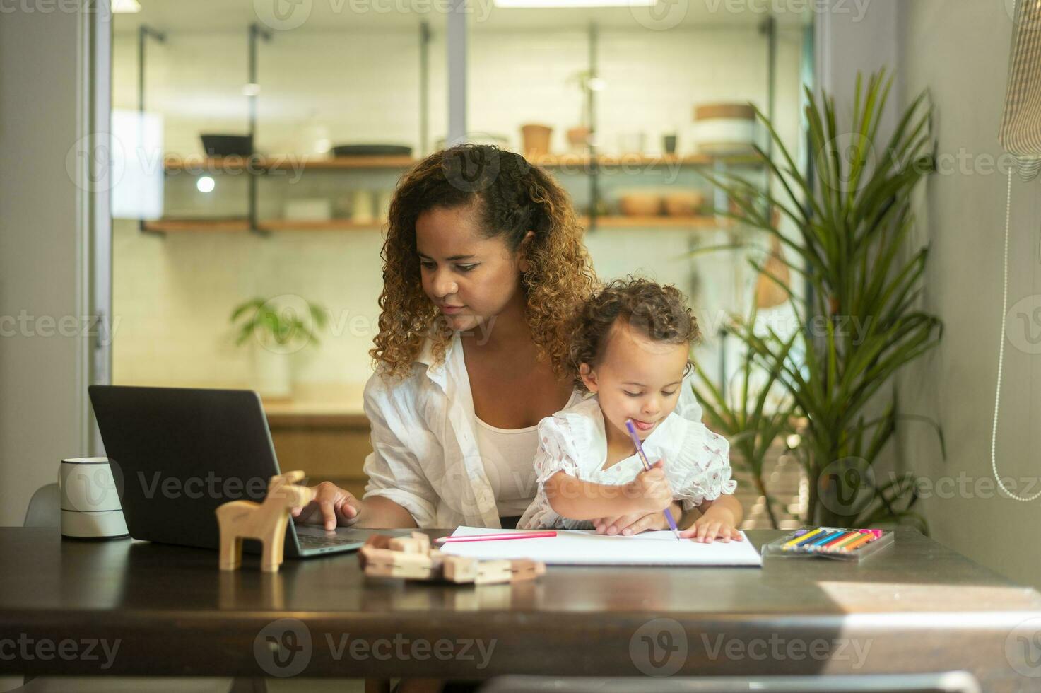 afrikanisch amerikanisch Mutter tragen beiläufig Kleider Arbeiten im ihr Zuhause während Tochter spielen oder Bildung foto