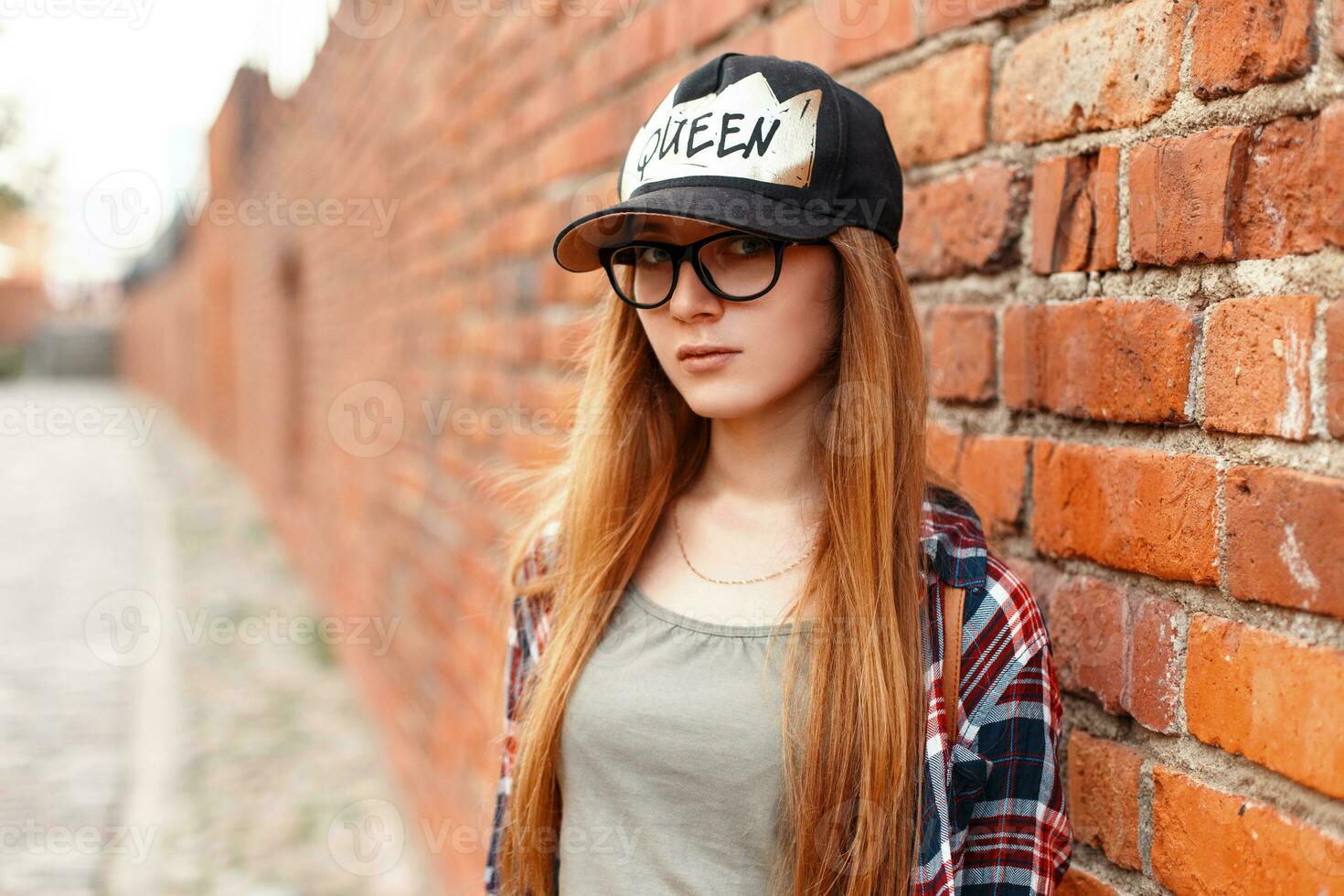 stilvoll ziemlich Hipster Frau mit Brille und ein Deckel in der Nähe von ein rot Backstein Mauer. foto