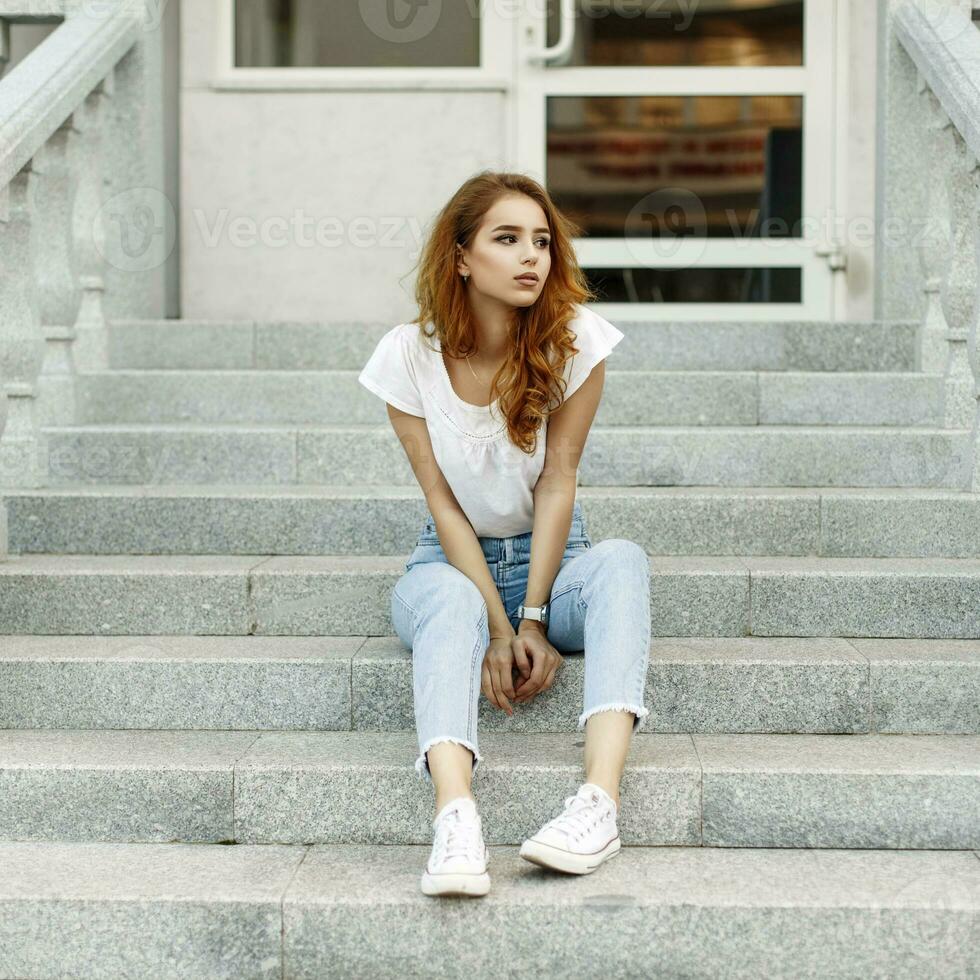 stilvoll jung Frau im ein Weiß Shirt, Jeans und Turnschuhe Sitzung auf das Stufen. foto