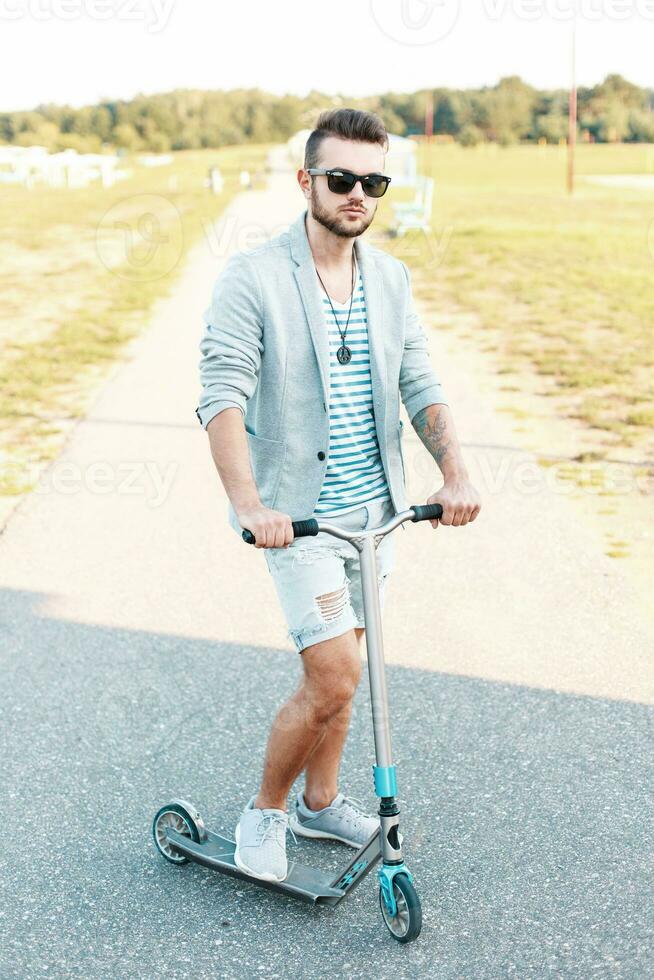 stilvoll gut aussehend Mann im modisch Kleider Reiten ein trete Roller auf das Strand foto