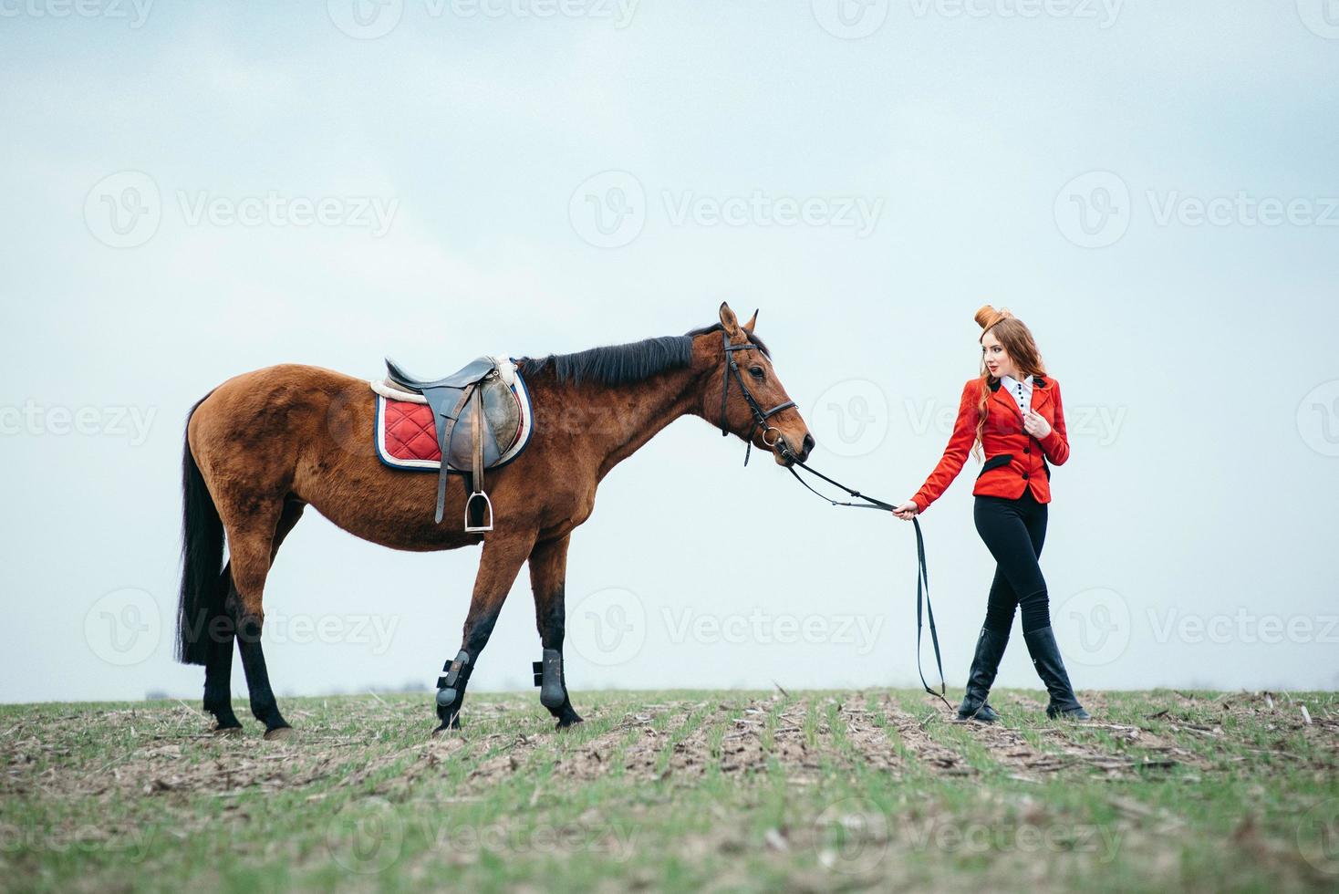rothaariges Jockey-Mädchen in einer roten Strickjacke und schwarzen hohen Stiefeln mit einem Pferd foto