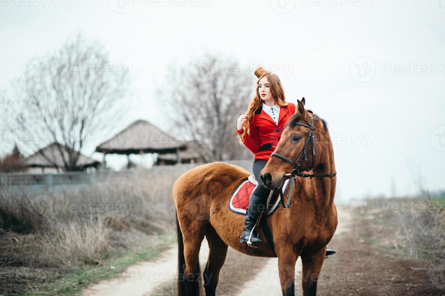 rothaariges Jockey-Mädchen in einer roten Strickjacke und schwarzen hohen Stiefeln mit einem Pferd foto