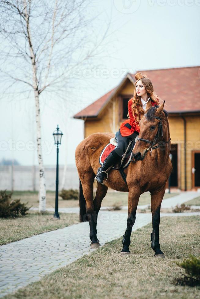 rothaariges Jockey-Mädchen in einer roten Strickjacke und schwarzen hohen Stiefeln mit einem Pferd foto