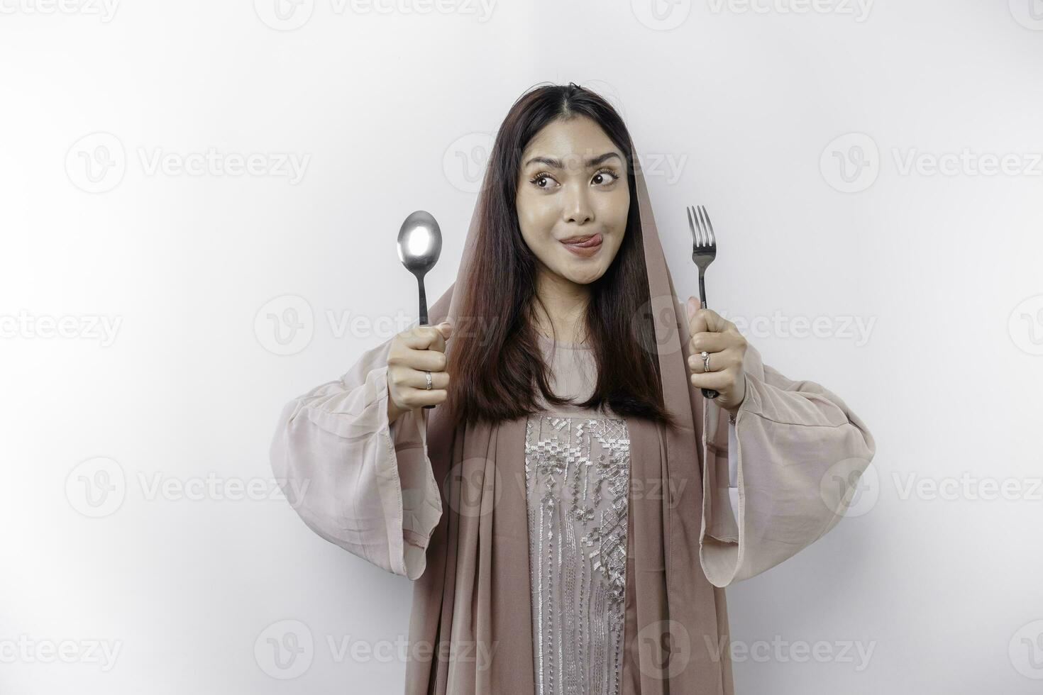 ein asiatisch Muslim Frau ist Fasten und hungrig und halten Besteck während suchen beiseite Denken Über Was zu Essen foto