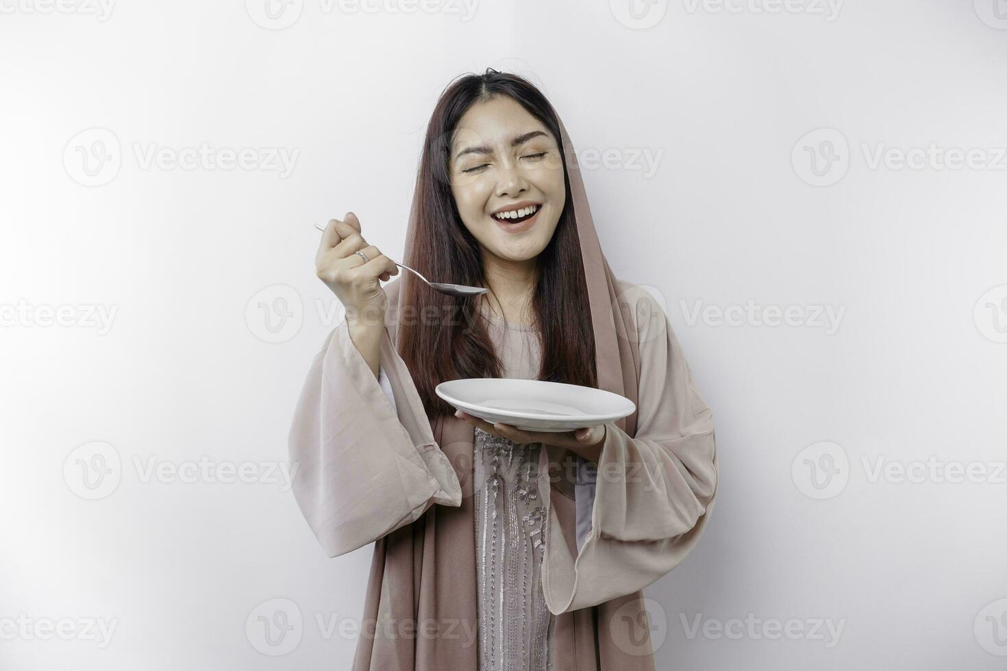 ein lächelnd asiatisch Muslim Frau ist Fasten und hungrig und halten und zeigen zu ein Teller foto