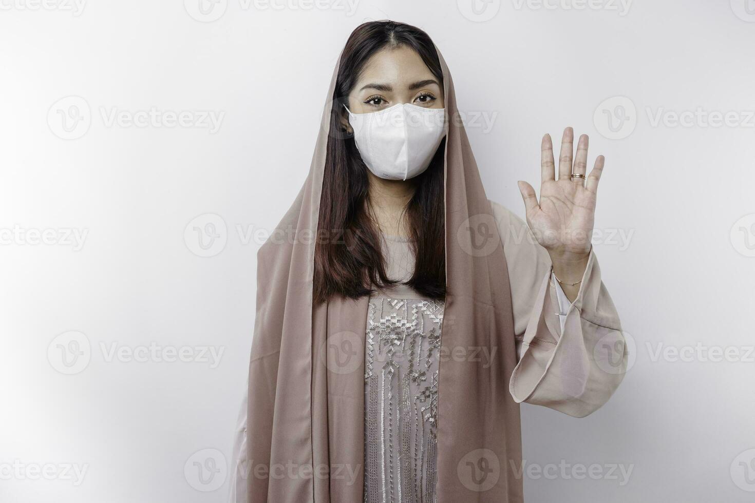 jung asiatisch Muslim Frau tragen ein Maske und Hijab zeigt an ihr Palme oder halt Geste, covid Verhütung Konzept foto
