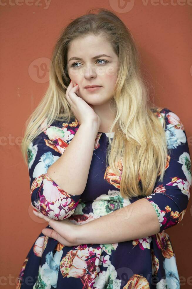 ein traurig blond Mädchen mit Blau Augen im ein Blau Kleid mit ein Blumen- drucken steht in der Nähe von ein Burgund Mauer. foto