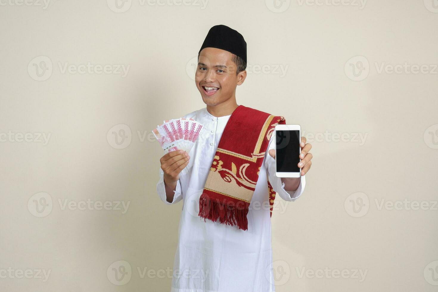 Porträt von attraktiv asiatisch Muslim Mann im Weiß Hemd mit Schädeldecke zeigen einer hundert tausend Rupiah während zeigen leer Bildschirm Handy, Mobiltelefon Telefon. finanziell und Einkaufen Konzept. isoliert Bild auf grau foto