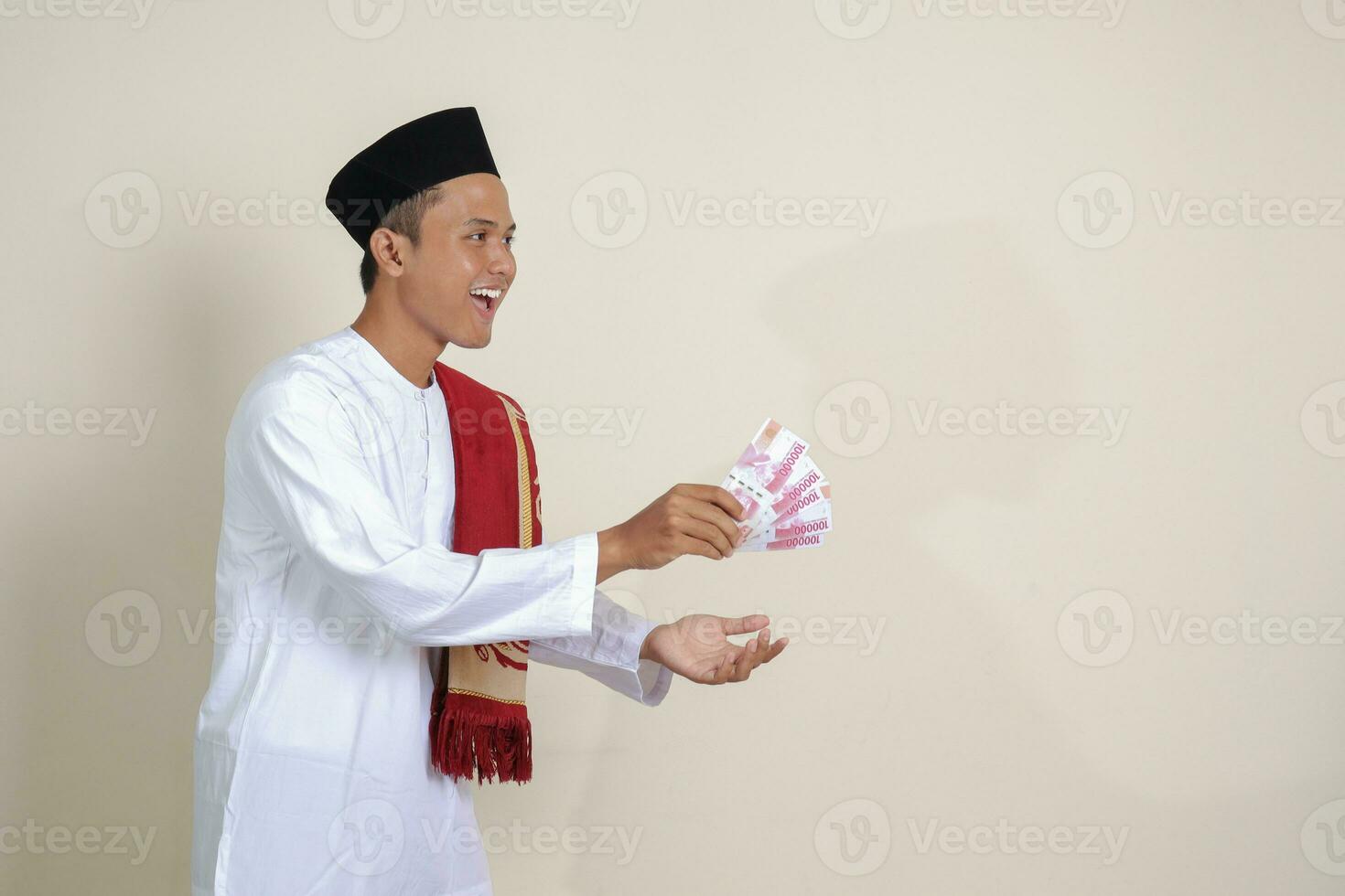 Porträt von attraktiv asiatisch Muslim Mann im Weiß Hemd mit Schädeldecke geben einer hundert tausend Rupiah zum Wohltätigkeit. isoliert Bild auf grau Hintergrund foto