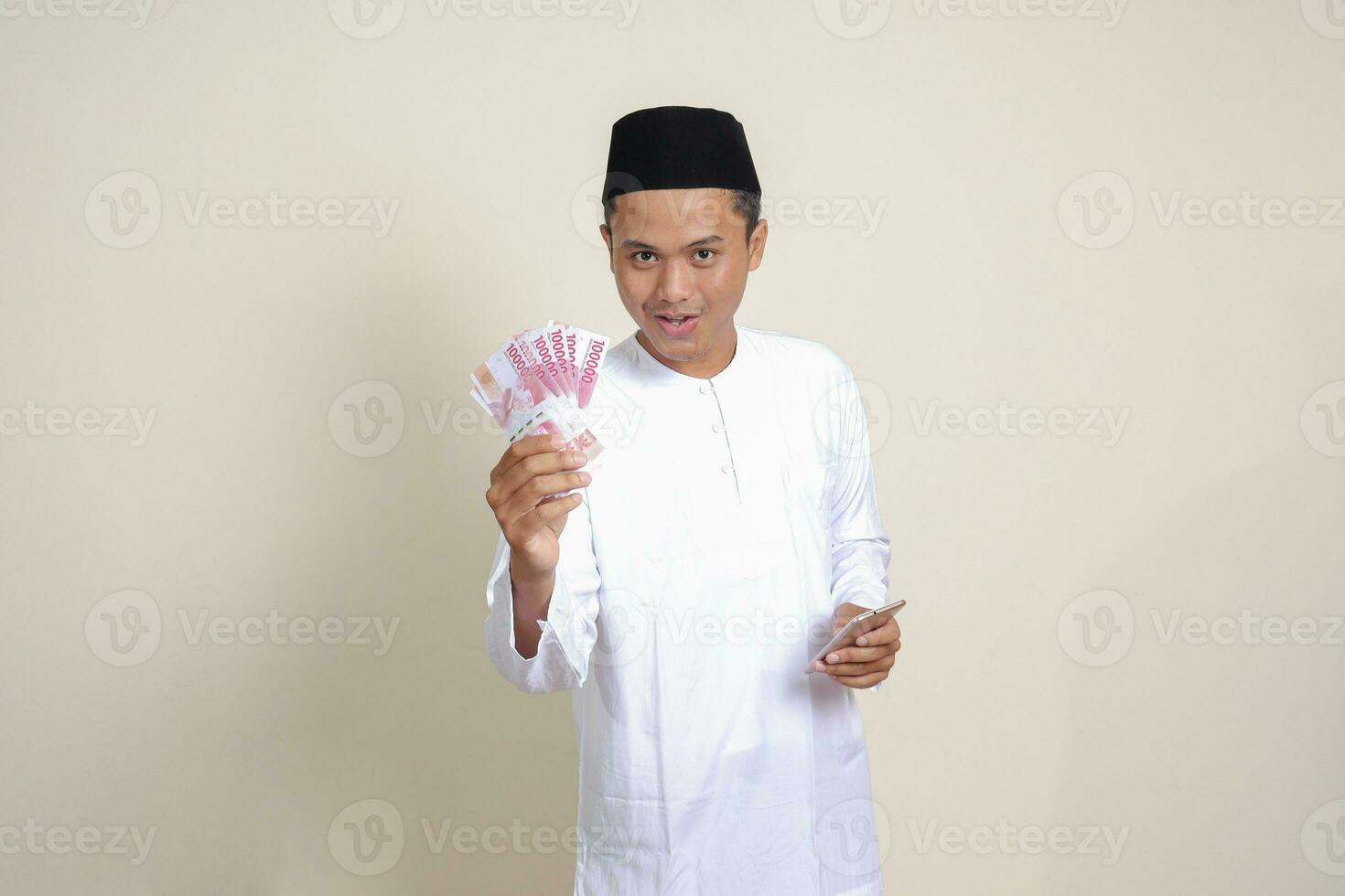 Porträt von attraktiv asiatisch Muslim Mann im Weiß Hemd zeigen einer hundert tausend Rupiah während mit Handy, Mobiltelefon Telefon. finanziell und Ersparnisse Konzept. isoliert Bild auf grau Hintergrund foto