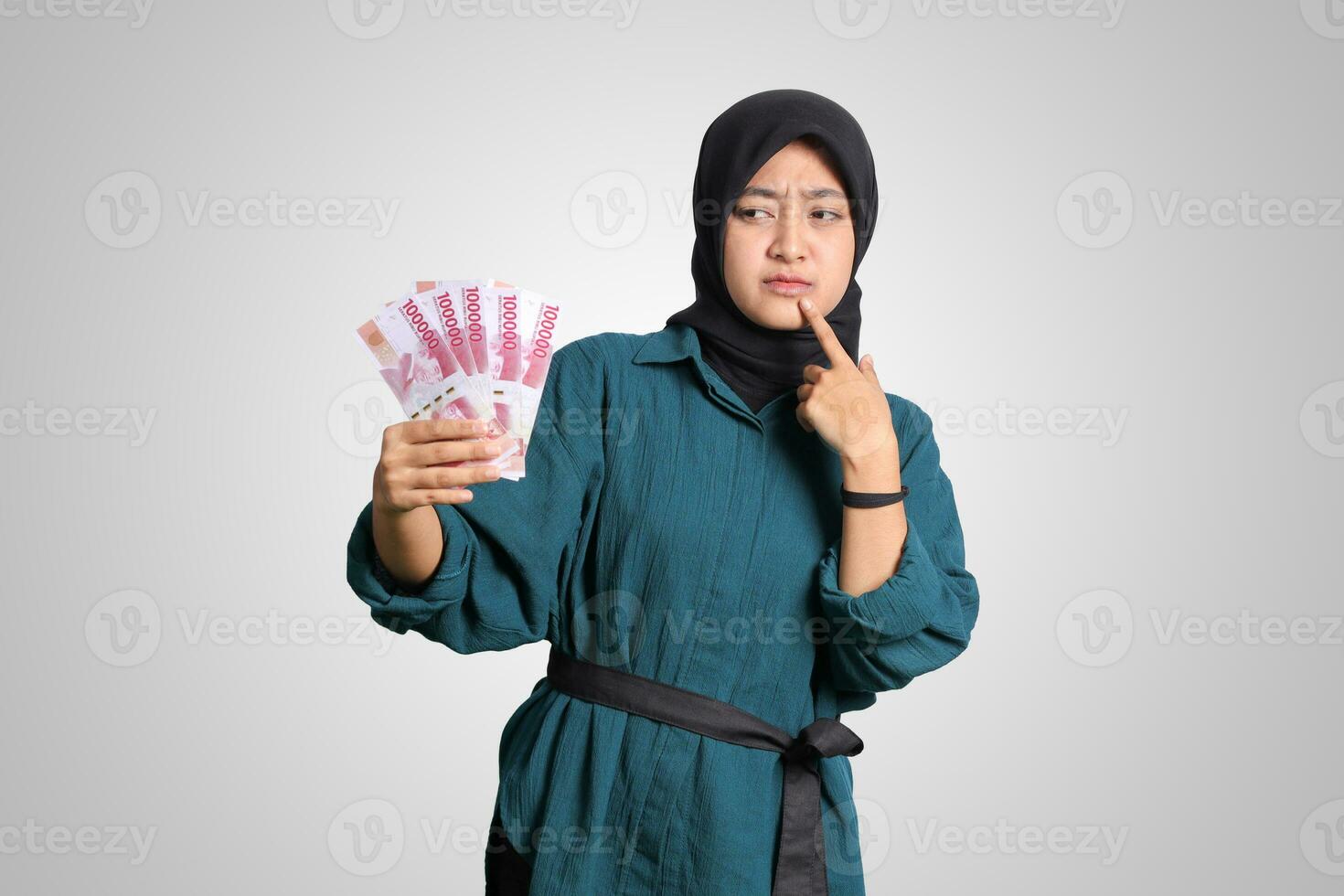 Porträt von verwirrt asiatisch Hijab Frau im beiläufig Outfit zeigen einer hundert tausend Rupiah während Denken mit Hand auf kinn. finanziell und Ersparnisse Konzept. isoliert Bild auf Weiß Hintergrund foto