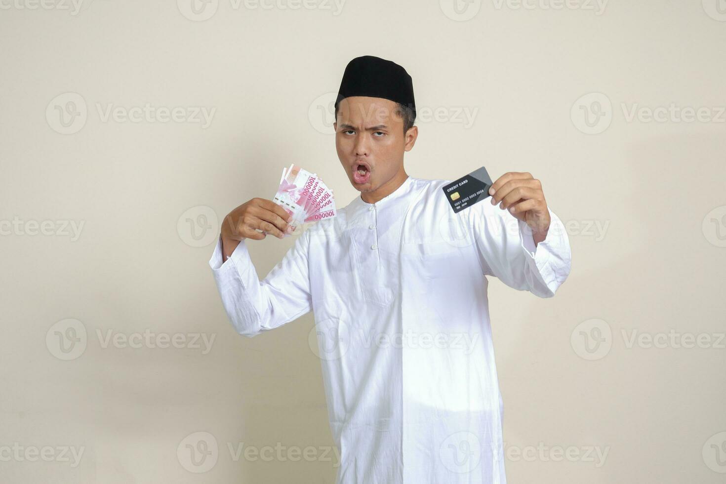 Porträt von attraktiv asiatisch Muslim Mann im Weiß Hemd mit Schädeldecke halten einer hundert tausend Rupiah und präsentieren Anerkennung Karte. isoliert Bild auf grau Hintergrund foto