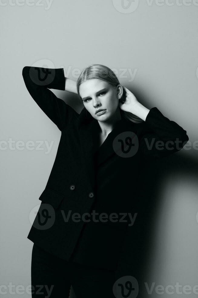 ein auffällig blond Mädchen im ein klassisch passen hält ihr Haar mit ihr Hände und posiert mögen ein Fachmann Modell- im ein Studio im schwarz und Weiß Stil. Mode Fotografie. Konzept zum Kleidung Shops foto