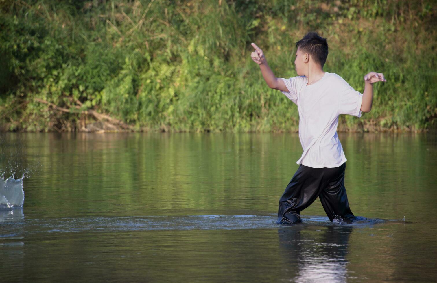asiatisch Junge im Weiß T-Shirt ist Ausgaben seine Freizeit durch Tauchen, Baden, werfen Felsen und fangen Fisch im das Fluss glücklich, Hobby und Glück von Kinder Konzept, im Bewegung. foto