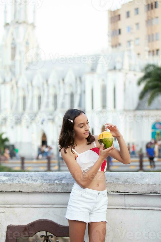 schön Tourist Mädchen Essen Mango beim das Ortiz Brücke mit la Ermita Kirche auf Hintergrund im das Stadt von cali im Kolumbien foto