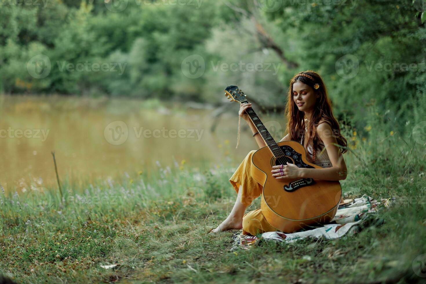ein Hippie Frau spielen ihr Gitarre lächelt und singt Lieder im Natur Sitzung auf ein Plaid im das Abend im das Sonnenuntergang Sonnenlicht. ein Lebensstil im Harmonie mit das Körper und Natur foto