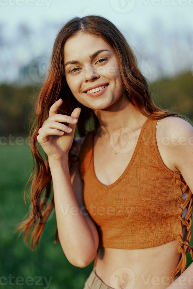 komisch jung rothaarig Frau im ein Orange oben lächelnd gegen das Grün Gras von Natur im das Sonnenuntergang Licht foto