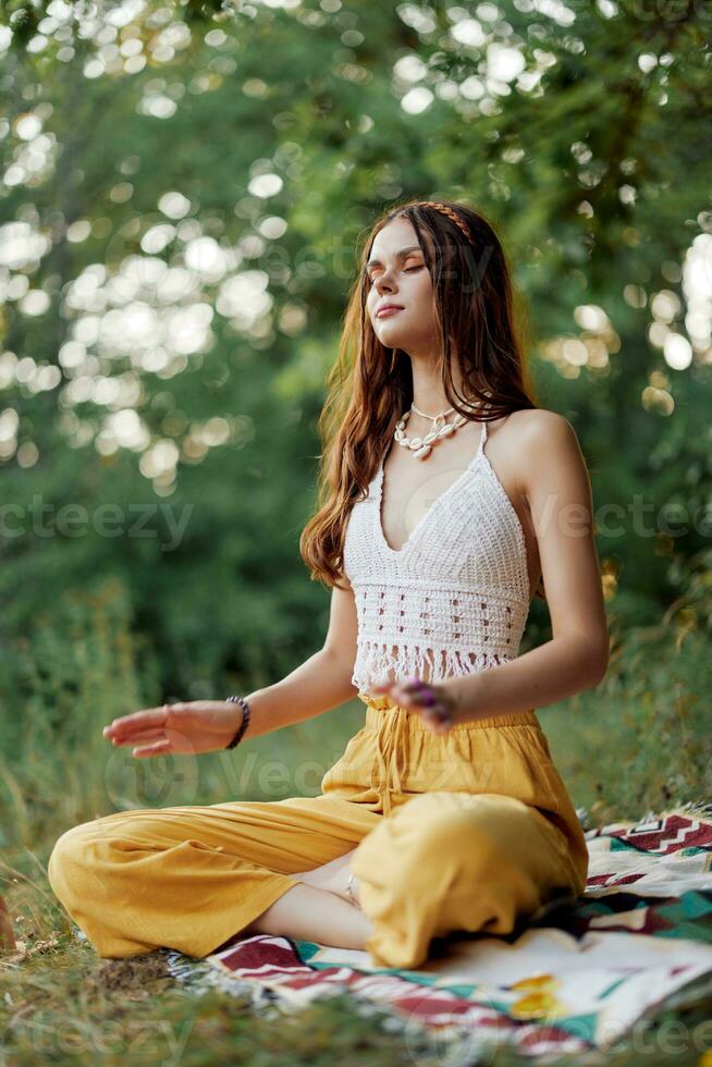ein jung Hippie Frau meditiert im Natur im das Park, Sitzung im ein Lotus Position auf ihr bunt Plaid und genießen Harmonie mit das Welt im Öko-Kleidung foto