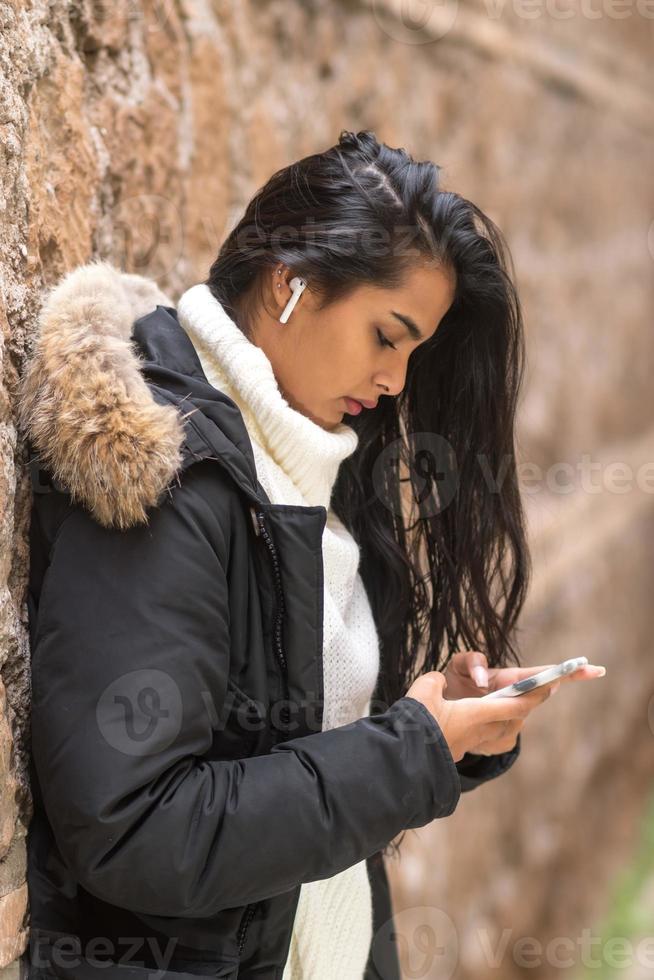 schöne brünette junge Frau in der Winterkleidung, die ihr Smartphone benutzt, das Musik mit Kopfhörern hört foto