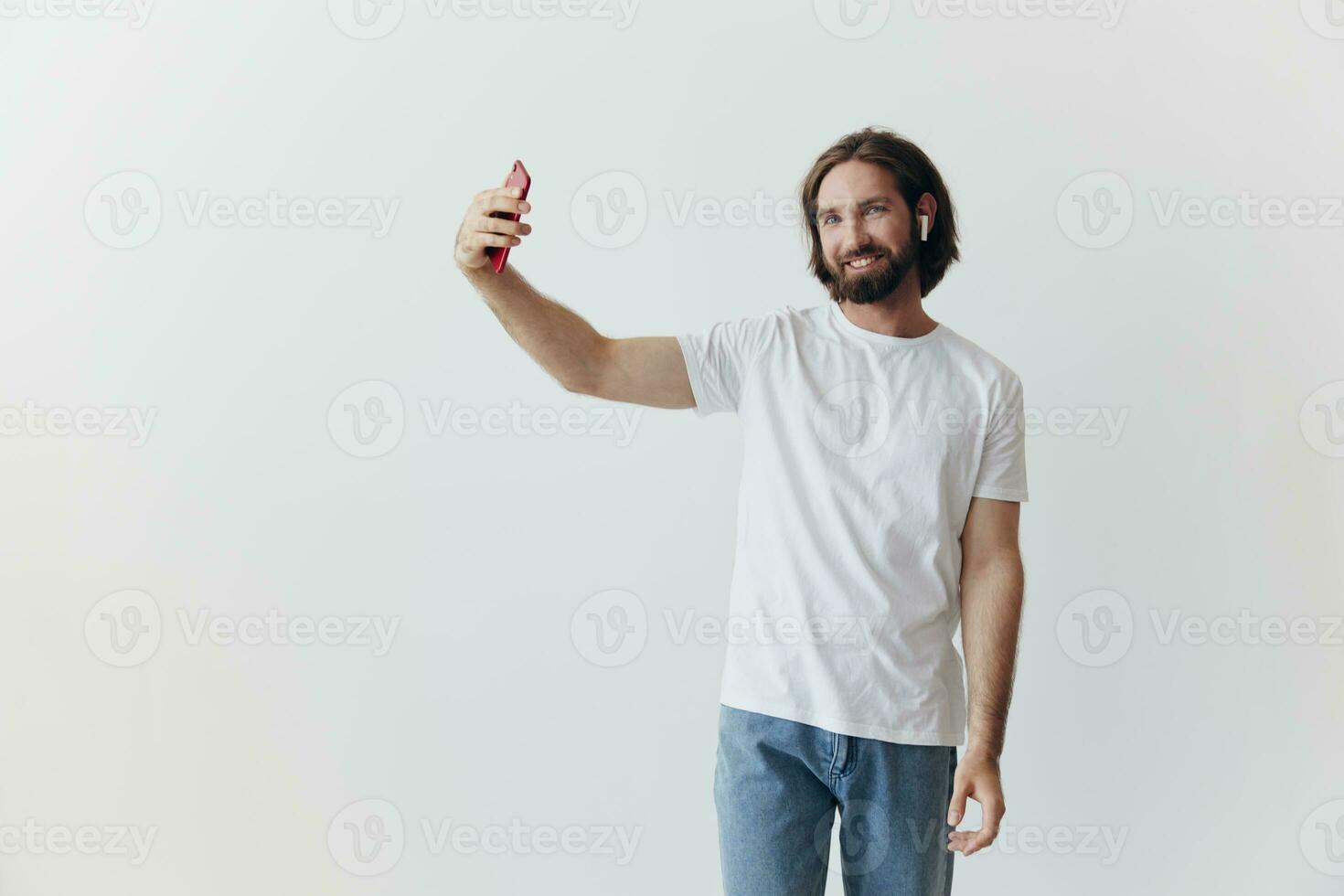 ein männlich Blogger Videoaufnahmen selbst auf seine Telefon und chatten mit Menschen online mit ein Lächeln im ein Weiß T-Shirt gegen ein Weiß Mauer foto