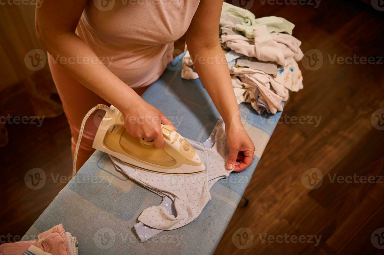 Overhead Sicht. Nahansicht schwanger Frau im dritte Semester von Schwangerschaft Bügeln Baby Bodysuit mit elektrisch Dampf Eisen foto