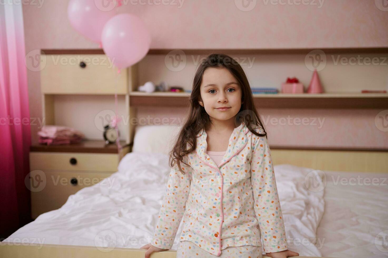 Porträt von selbstbewusst wenig Mädchen mit schön lange Haar, im Pyjama Sitzung auf das Bett im ein Glanz Rosa Schlafzimmer foto