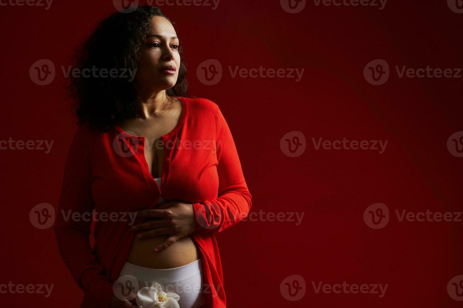 attraktiv schwanger Frau gekleidet im rot tragen, Stehen nackt Bauch mit ein Weiß Orchidee im Hände, nachdenklich suchen Weg foto