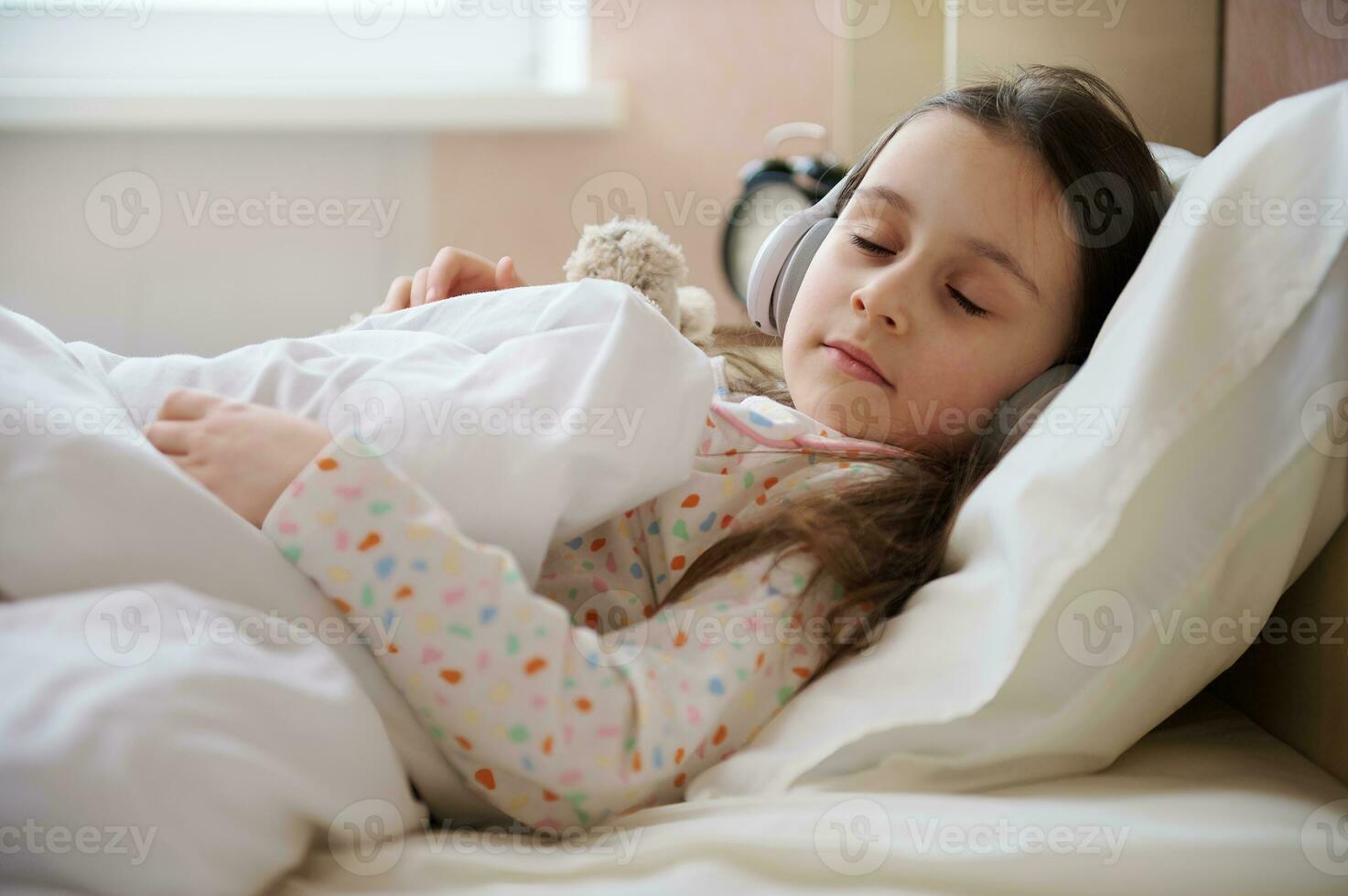 bezaubernd Kind Mädchen im Kopfhörer, Nickerchen machen im ein Weiß Schlafzimmer, fallen schlafend zu beruhigend Musik- auf ihr komfortabel Bett foto