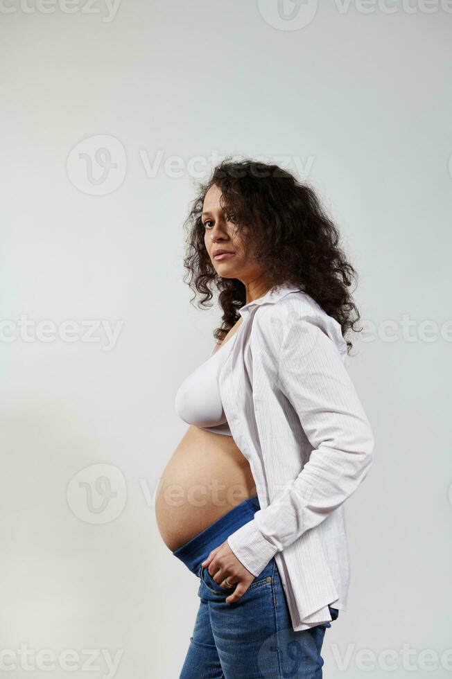 Vertikale Porträt von lockig behaart schwanger Frau, posieren nackt Bauch auf isoliert Weiß Hintergrund. glücklich Schwangerschaft 6 Monat foto