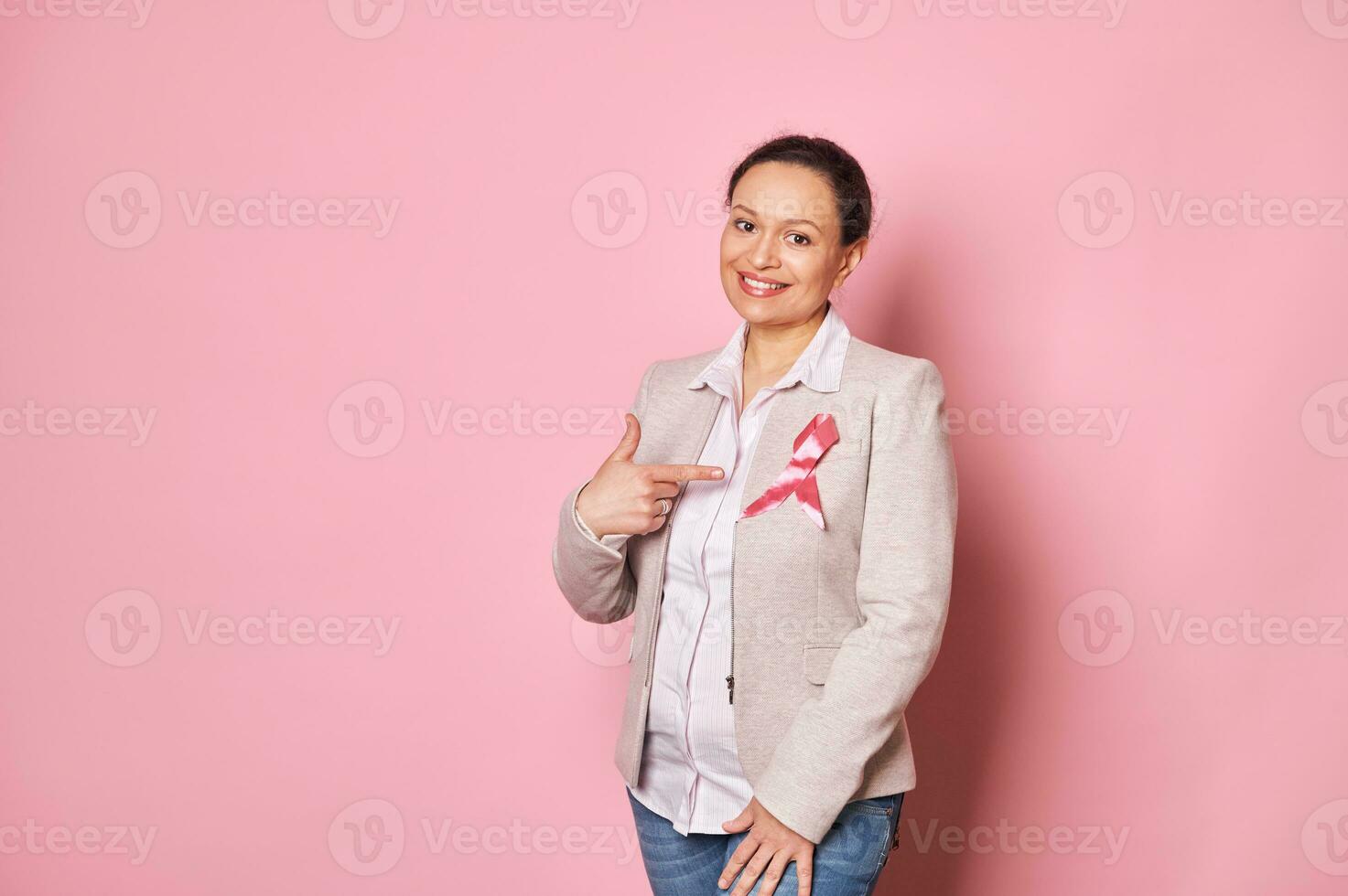positiv multiethnisch Frau, Stehen Über Rosa Hintergrund und zeigen ihr Finger beim ein Rosa Satin- Band auf ihr Jacke foto