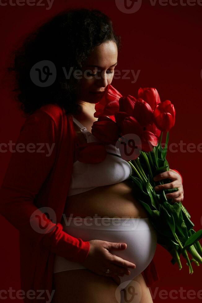 schwanger Frau, erwartungsvoll Mutter schnüffeln ein Strauß von rot Tulpen, sanft streicheln ihr Bauch, auf isoliert Hintergrund foto