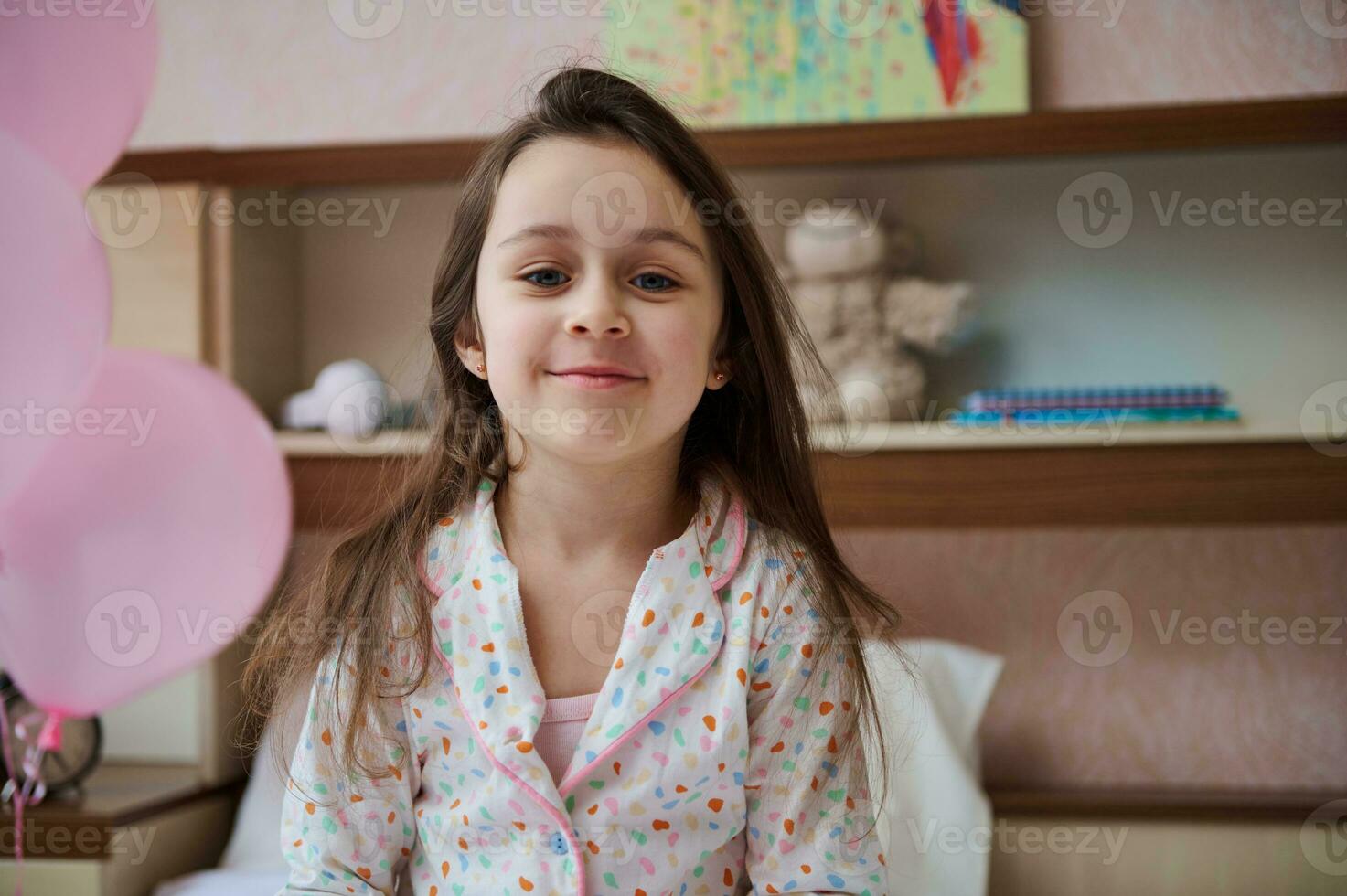 kaukasisch wenig 5 Jahre alt Kind Mädchen im Pyjama, lächelnd suchen beim Kamera, Sitzung auf das Bett im ihr Glanz Schlafzimmer foto