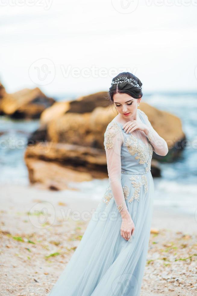 Braut in einem Blaulichtkleid, das entlang des Ozeans geht foto