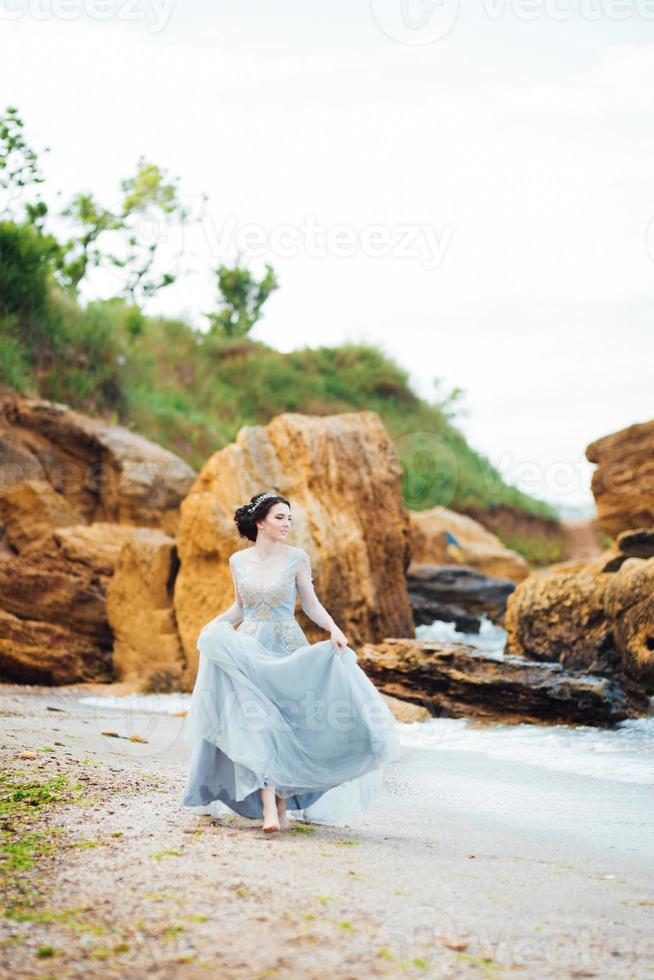 Braut in einem Blaulichtkleid, das entlang des Ozeans geht foto