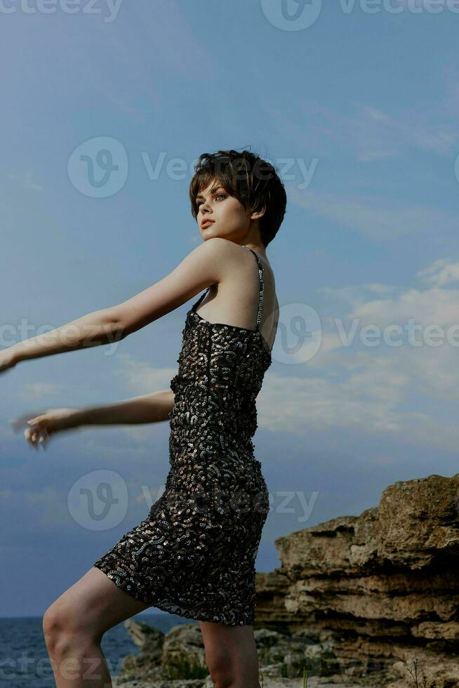 Frau mit kurz Haar im ein glänzend Kleid posieren gegen das Hintergrund von ein Felsen foto