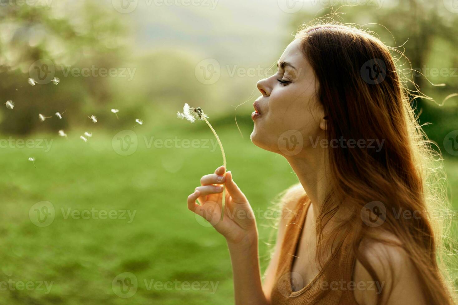 Frau mit ein Löwenzahn Blume im ihr Hände lächelnd und weht auf es gegen ein Hintergrund von Sommer- Grün und Sonnenlicht foto