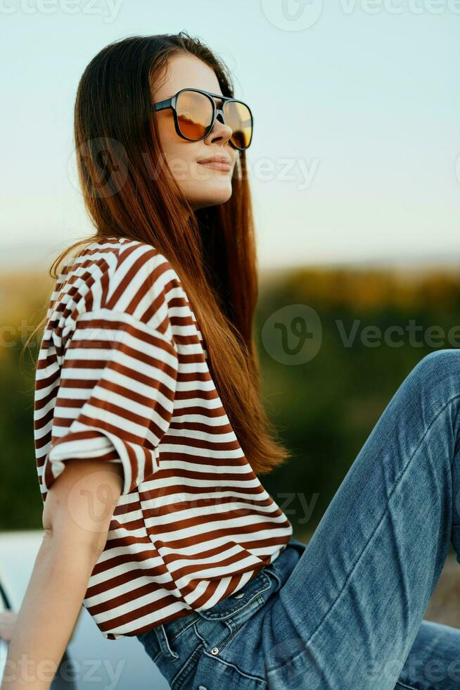 ein Mode Frau im stilvoll Sonnenbrille, ein gestreift T-Shirt und Jeans sitzt auf das Kofferraum von ein Auto und sieht aus beim das schön Natur von Herbst. Reise Lebensstil foto