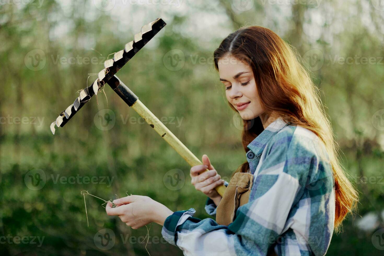 Frau schön lächelnd Farmer im Arbeit Kleider und Schürze Arbeiten draußen im Natur und halten ein Rechen zu versammeln Gras foto