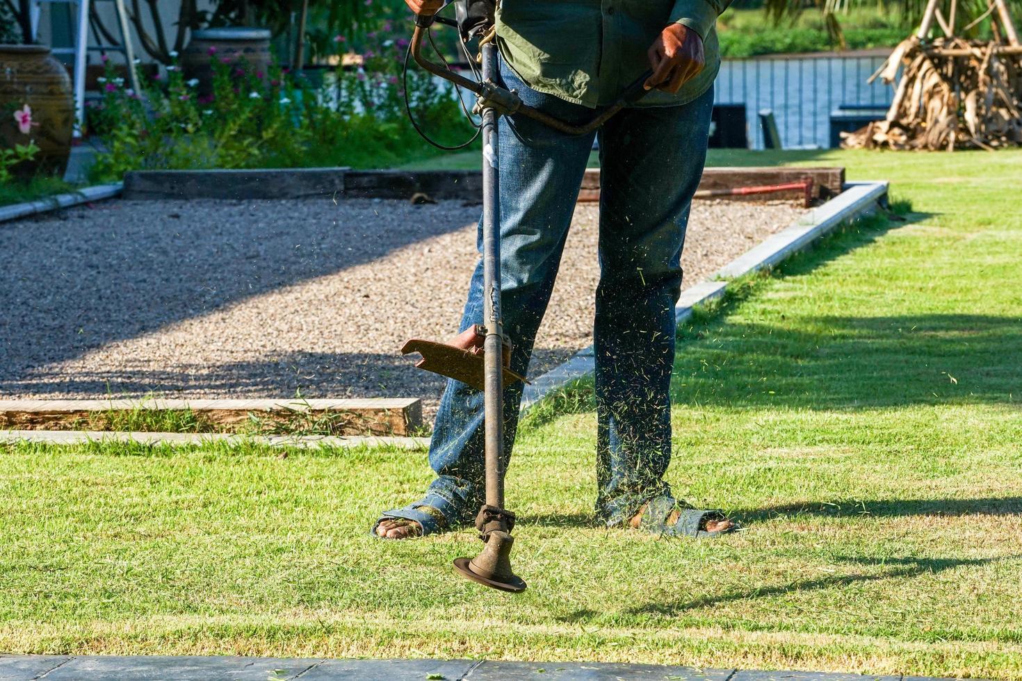 Schließen Sie die Beine eines Gärtners, der an einem sonnigen Tag das Gras in einem Garten im Freien mit Mähwerkzeugen mäht foto