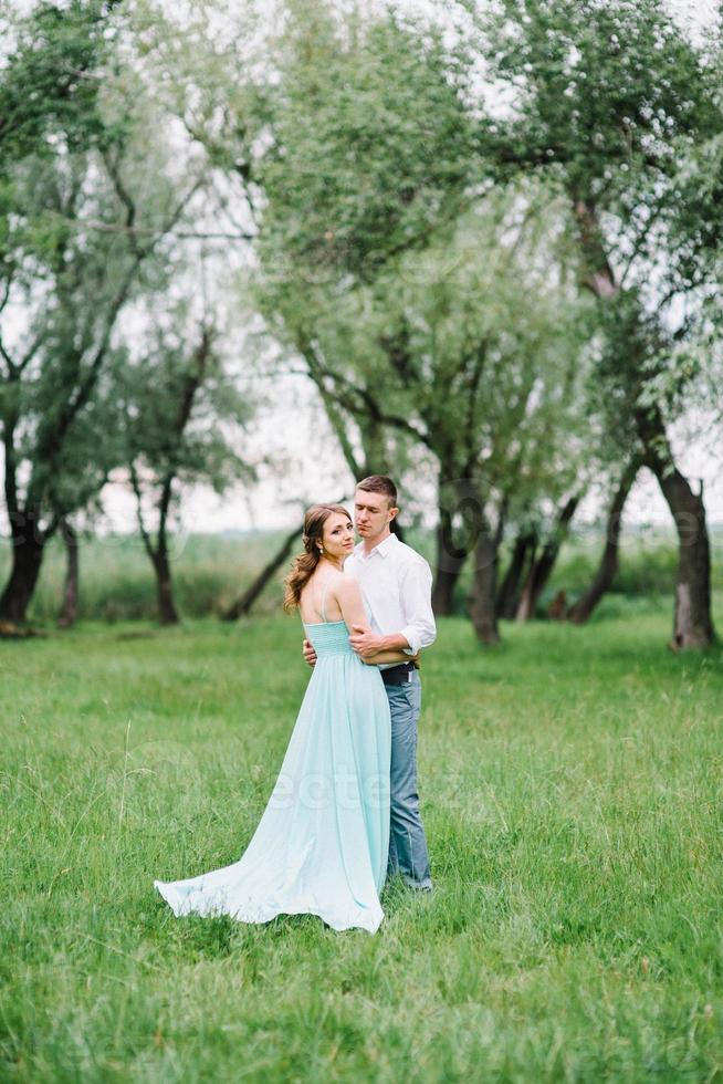 Ein glücklicher Kerl in einem weißen Hemd und ein Mädchen in einem türkisfarbenen Kleid gehen im Waldpark spazieren foto