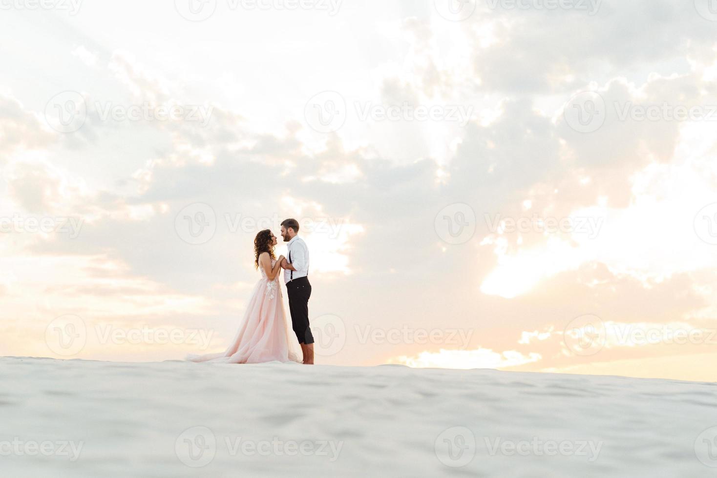 Ein junges Paar, ein Mann in einer schwarzen Reithose und ein Mädchen in einem rosa Kleid, gehen den weißen Sand entlang foto