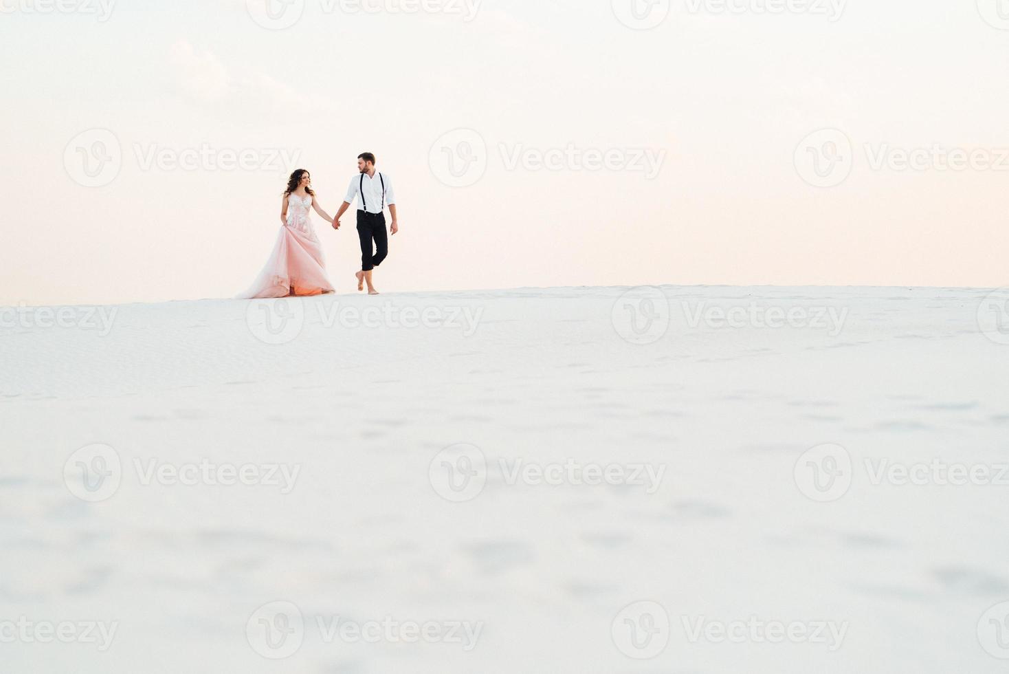 Ein junges Paar, ein Mann in einer schwarzen Reithose und ein Mädchen in einem rosa Kleid, gehen den weißen Sand entlang foto