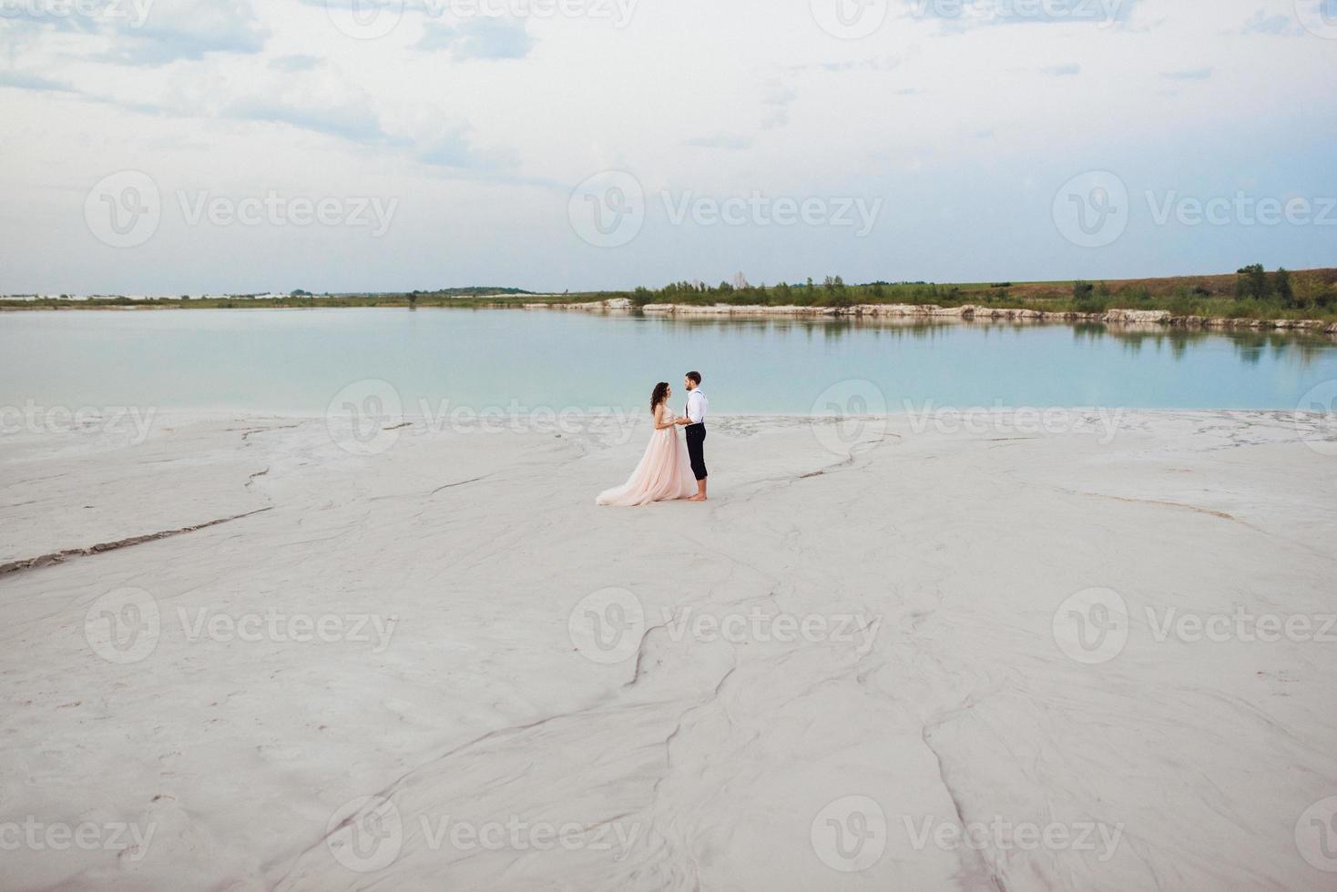 Ein junges Paar, ein Mann in einer schwarzen Reithose und ein Mädchen in einem rosa Kleid, gehen den weißen Sand entlang foto