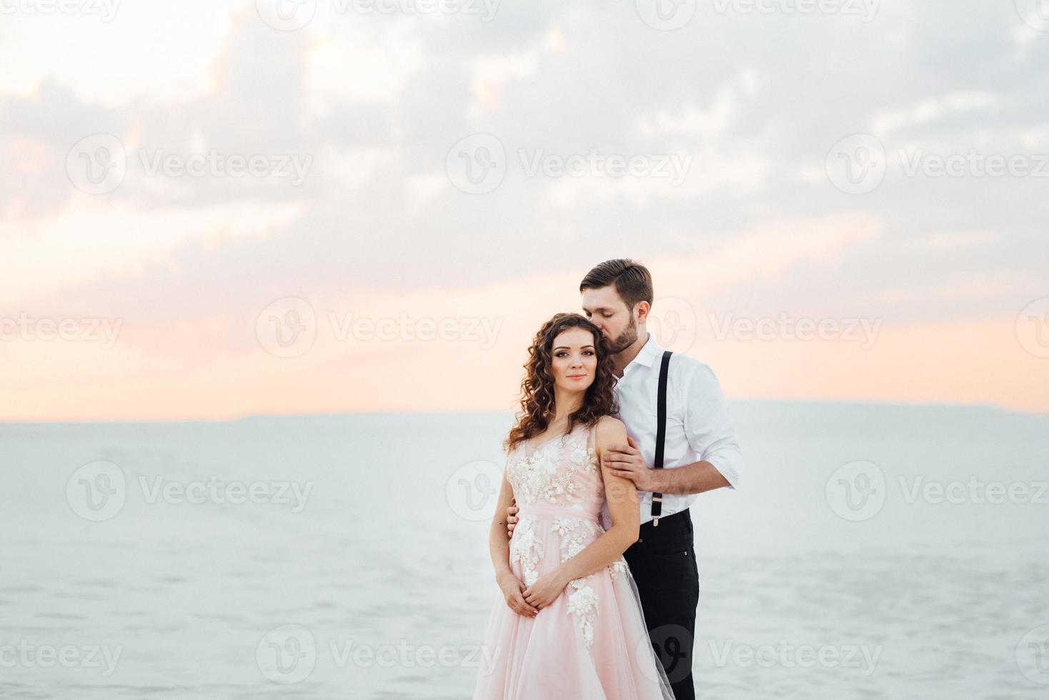 Ein junges Paar, ein Mann in einer schwarzen Reithose und ein Mädchen in einem rosa Kleid, gehen den weißen Sand entlang foto