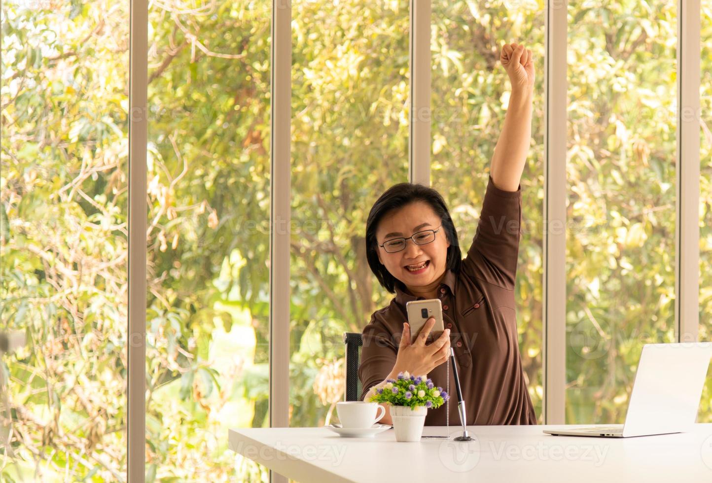 ältere asiatische Frau, die ein Mobiltelefon und einen Laptop im natürlichen Wohnzimmerhintergrund mit moderner Technologie verwendet foto