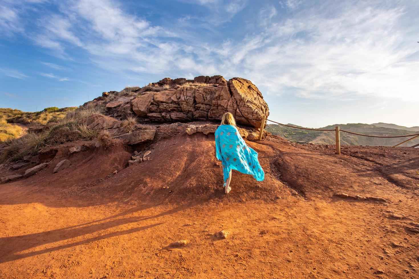 Weitwinkelbild mit dramatischem blauen Himmel und schöner Landschaftsansicht bei Sonnenuntergang der Frau mit einem blauen Schleier, der in Richtung roter Felsen in Menorca-Insel geht foto