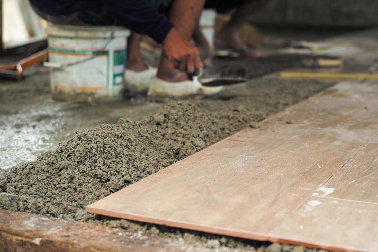 Selektiver Fokus auf die Textur von gemischtem Zement und schmutziger Oberfläche von Bodenfliesen im im Bau befindlichen Haus mit unscharfem Arbeiter im Hintergrund foto