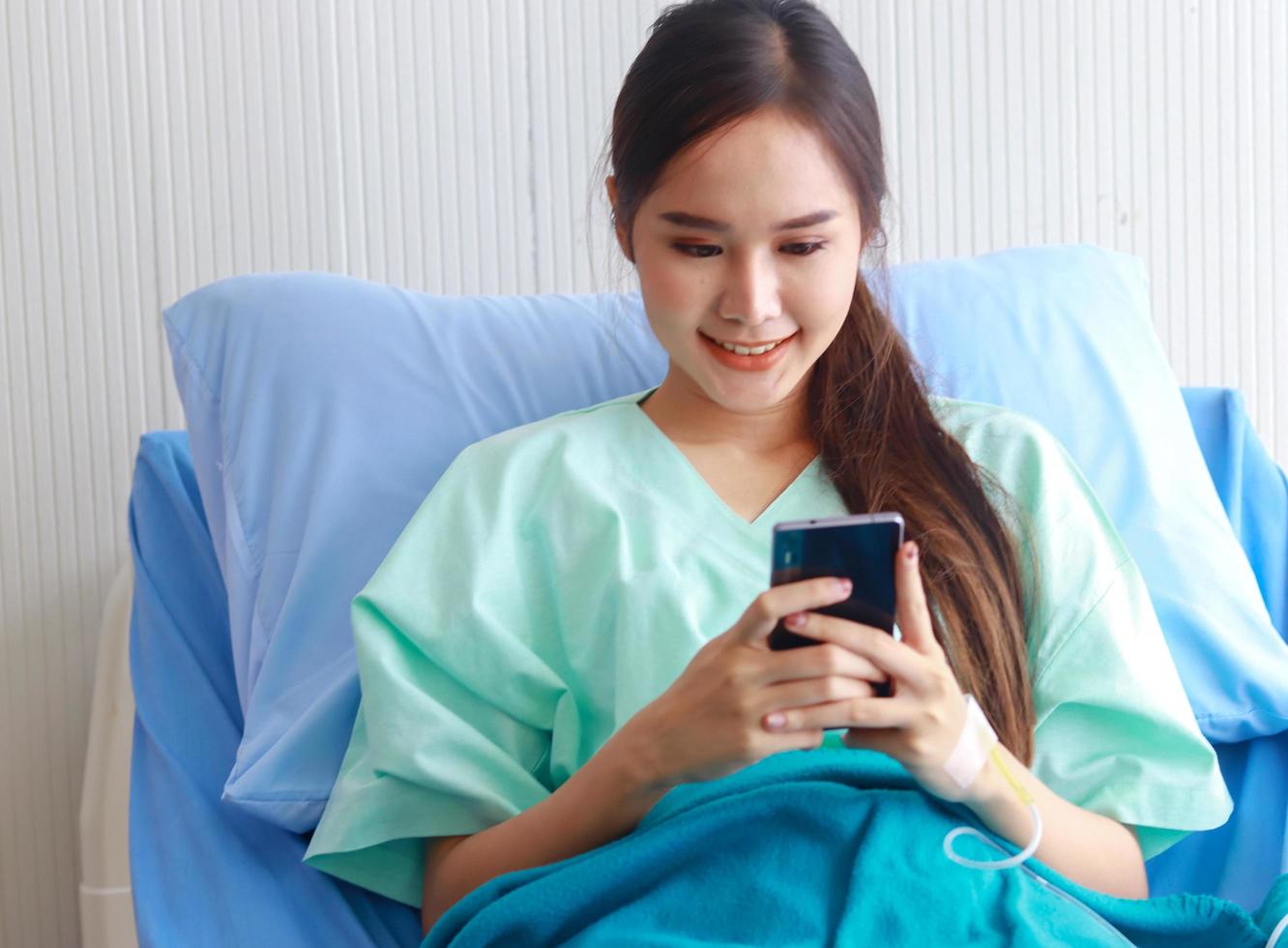 schöne asiatische Frau, die auf einem Krankenhausbett sitzt und mit ihrem Smartphone spielt foto