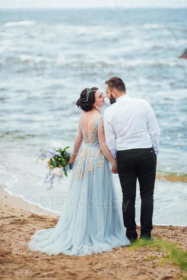 Braut in einem blauen Kleid mit Bräutigam, der entlang der Ozeanküste geht foto